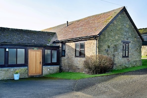 Un bâtiment en pierre avec une porte et des fenêtres en bois, une allée gravillonnée et un petit arbuste devant.