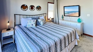 A neatly made bed with a striped bedspread, a bedside table with a lamp, a dresser with a mirror, and a wall-mounted TV displaying a beach scene. - Blue Lagoon | Water Views + Walkable Beach Access (Topsail Beach)
