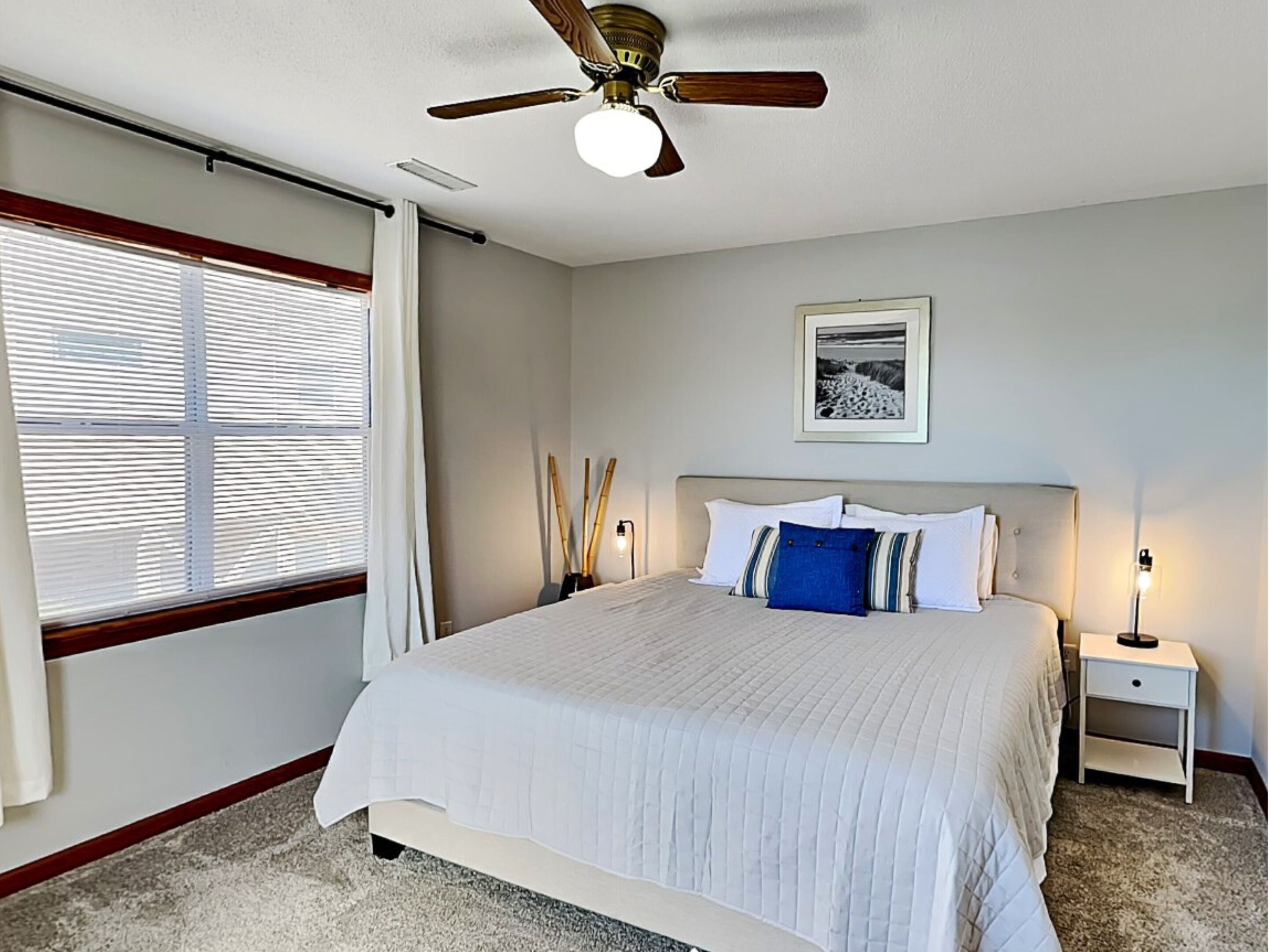 A bedroom with a bed, bedside tables, a ceiling fan, a window with blinds, and a framed picture on the wall.