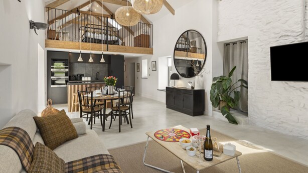 A spacious living area with a high ceiling, wooden beams, a dining table with chairs, a sofa, a coffee table with a bottle, and a large mirror. - Stylish barn conversion, sleeps 5, with hot tub (Carmarthen)