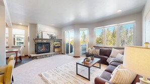A spacious living room with a stone fireplace, a sofa, a coffee table, and large windows offering a view of trees.