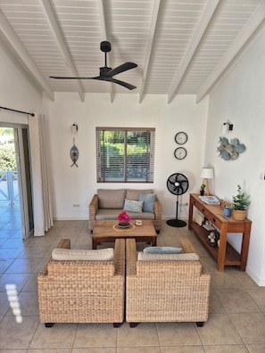 A living room with a sofa, two armchairs, a coffee table, a ceiling fan, a window with blinds, wall clocks, and a small table with decorative items.