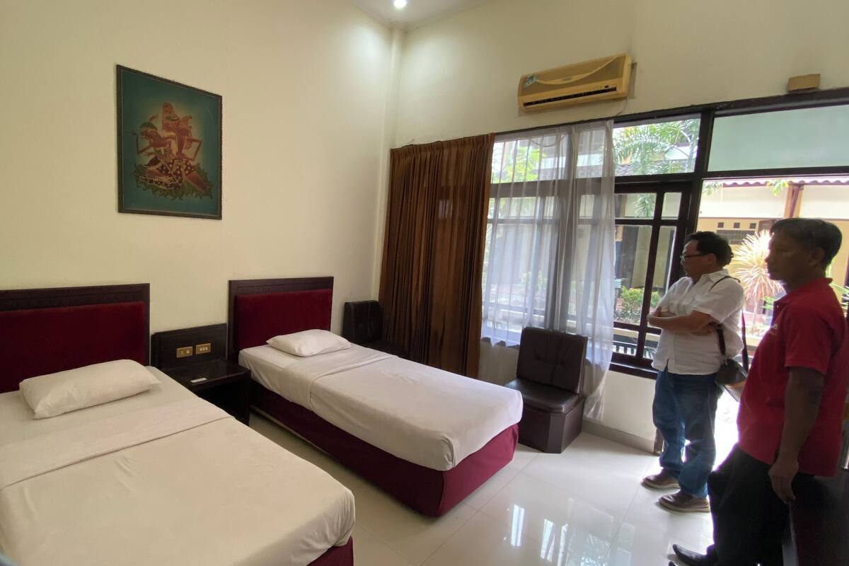A hotel room with two single beds, a large window with curtains, and a painting on the wall.