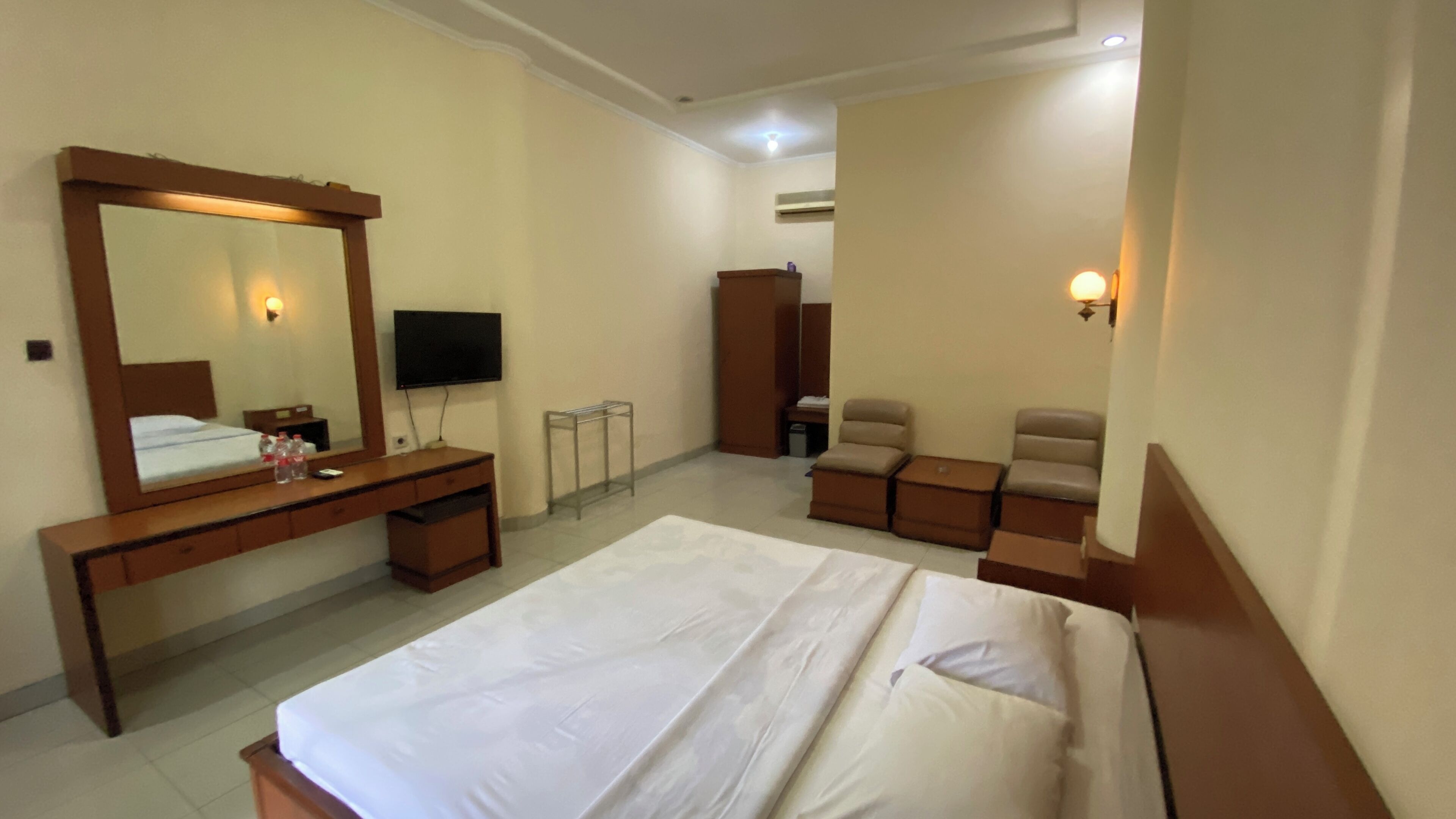 Una habitación de hotel con una cama grande, un aparador de madera, un televisor, un armario y una zona de estar con sillas y otomanas.