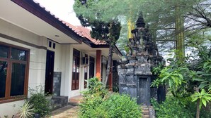A traditional Balinese temple with a stone structure, surrounded by lush greenery and a modern building with large windows and a tiled roof.