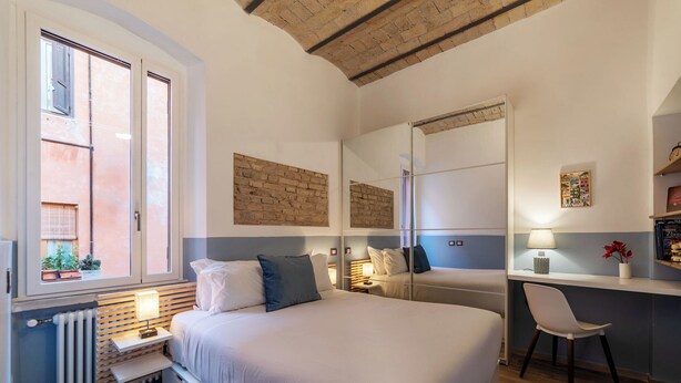 Ein Schlafzimmer mit Bett, Nachttischen, Schreibtisch, Stuhl, Heizkörper und einem Fenster mit Jalousien.