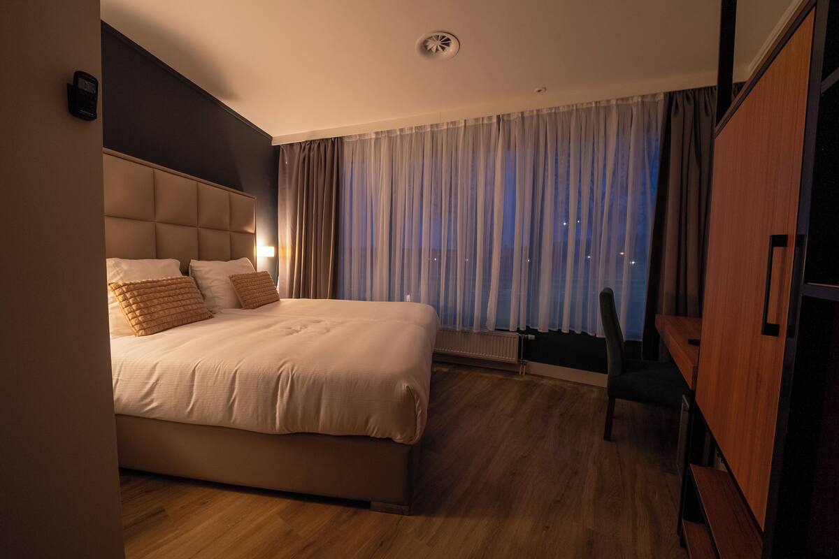 A modern hotel room with a large bed, a wooden floor, a desk, and a view of the city at night.