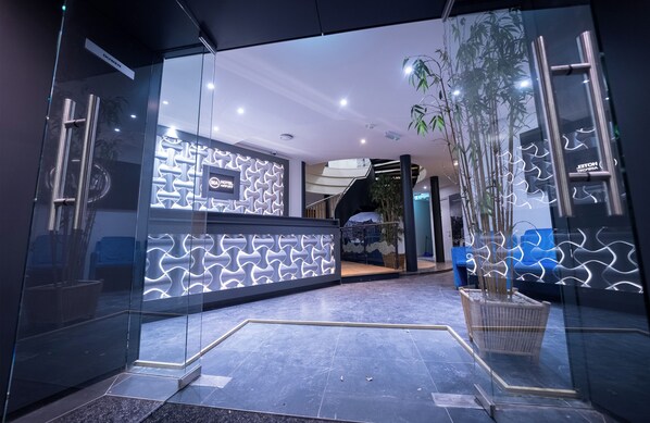 A modern reception area with a glass entrance, a reception desk with a geometric light pattern, and a staircase leading to an upper level. - Ma Airport Hotel (Hoofddorp)