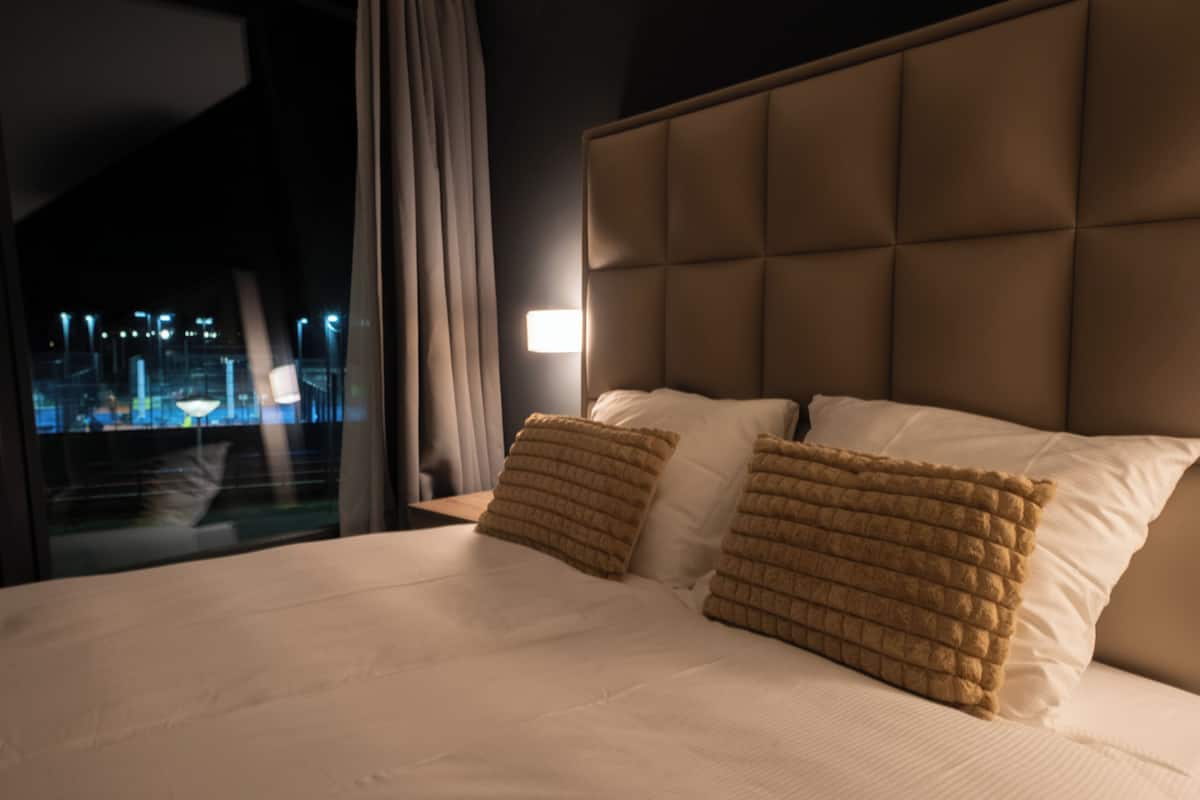 A hotel room with a large bed, a headboard, and a view of a cityscape at night.