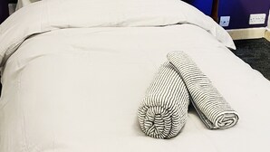 Ein ordentlich gemachtes Bett mit weißen Leinentüchern, einem gestreiften Kopfkissen und aufgerollten Handtüchern auf einem Nachttisch aus Holz neben einer violetten Wand.