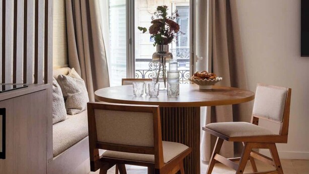 A dining area with a round table, two chairs, a vase with flowers, and a window with curtains.