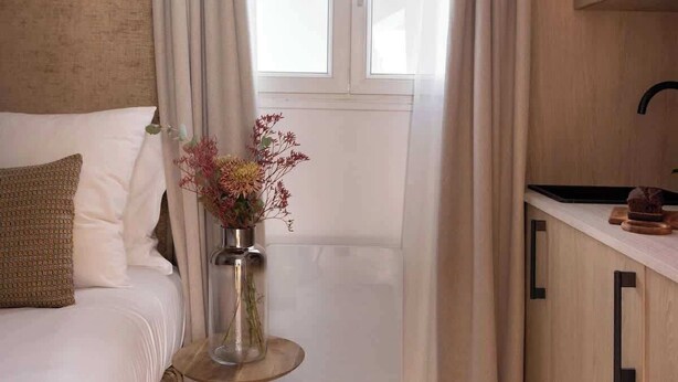 A bedroom with a bed, a bedside table with a vase of flowers, a window with curtains, and a kitchenette area. - Charming modern Studio with AC - Le Marais (Paris)