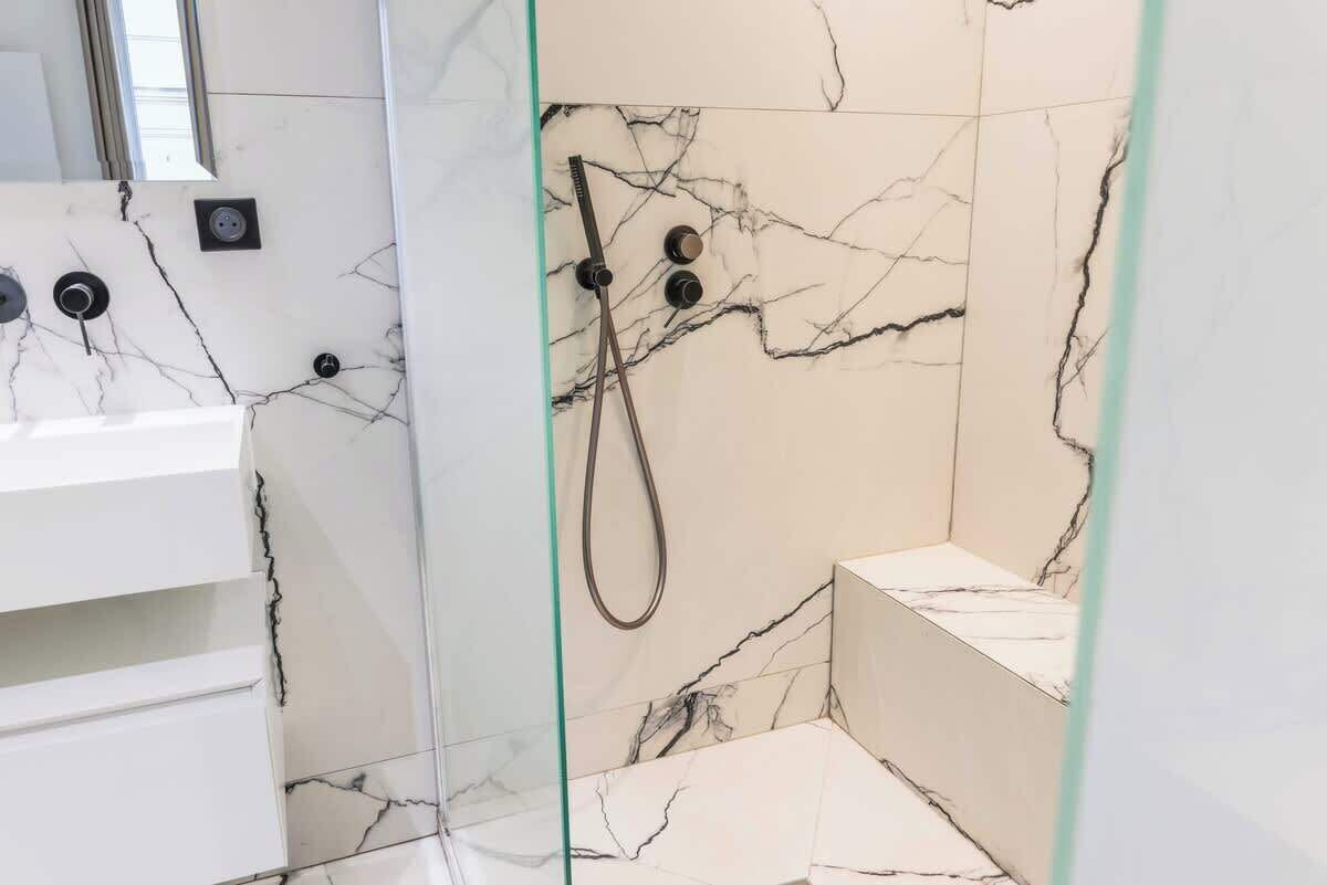 Un baño con pared de mármol, cabina de ducha de vidrio y lavamanos blanco.