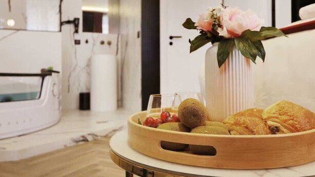 A modern bathroom with a white bathtub, a wooden tray with pastries and fruits, and a vase with flowers.