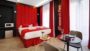 A hotel room with a bed, a dining table with chairs, a small kitchen area, and a neon sign on the wall.