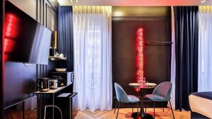 A modern hotel room with a flat-screen TV, a small kitchenette, a dining area with two chairs, and a large window with curtains.
