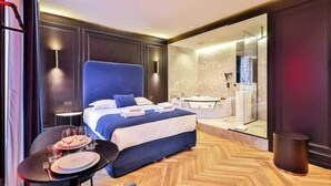 A modern hotel room with a large bed, a glass-enclosed bathroom, and a dining area with a table set for two.