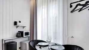 Habitación de hotel moderna y minimalista con zona de comedor que incluye una mesa redonda, dos sillas negras y un vaso de agua. Se observa una kitchenette compacta con microondas y cafetera, así como un altavoz negro en la pared.