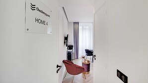 Una habitación de hotel moderna con un cartel de recepción, una silla roja, una mesita con un florero y vista del área de estar a través de una puerta.
