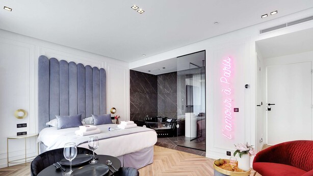 Una habitación de hotel moderna con una cama, una zona de comedor con mesa y sillas, un sillón rojo y un letrero de neón que dice 
