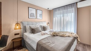 Una habitació d'hotel moderna amb un llit gran, una taula auxiliar, una cadira i una finestra amb cortines transparents.