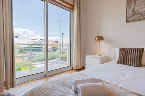 A bedroom with a large sliding glass door, a bed with white bedding, a bedside lamp, and a view of buildings and greenery outside. - 2 Bedroom Relax in Alvor by Innkeeper (Portimão)