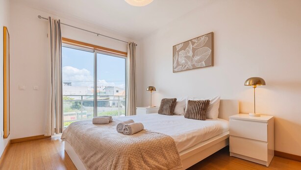 Een slaapkamer met een groot bed, twee nachtkastjes, een raam met gordijnen, een aan de muur hangend kunstwerk en uitzicht op gebouwen.