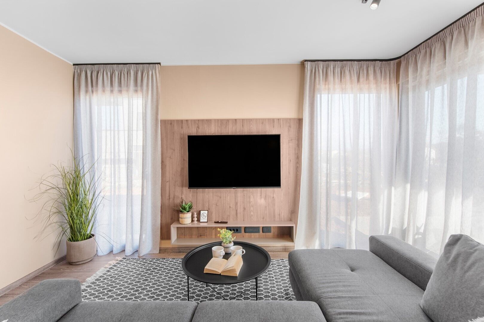 Una sala de estar moderna con un televisor de pantalla plana montado en una pared de madera, sofás grises, una mesa de centro con un libro y tazas, y una planta en maceta.