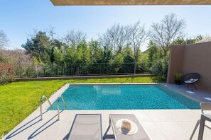 A rectangular swimming pool with clear blue water, surrounded by a grassy area and trees. There are lounge chairs and a small table with a potted plant nearby. - Ground-Floor Apartment with Private Pool and Terrace in Istria, 150 Meters from the Sea (Brtonigla)