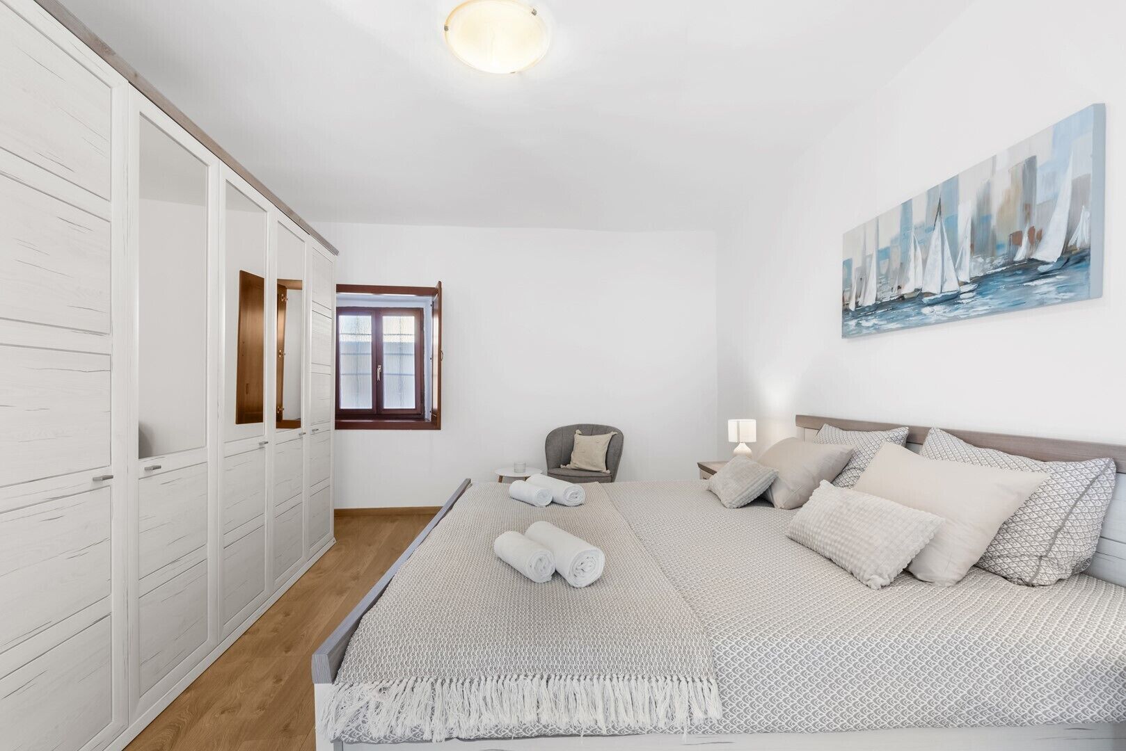 Ein Schlafzimmer mit einem großen Bett, einem Kleiderschrank, einem Fenster, einem Sessel und einem Bild an der Wand.