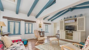 A living room with wooden floors, blue beams, a sofa, a chair, a TV stand, and a patterned rug.