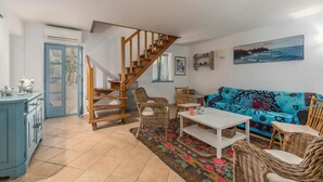 A living room with a wooden staircase, a blue sofa, wicker chairs, a white table, and a colorful rug.