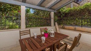 A wooden table with chairs and a pink flower pot, set under a pergola with a view of greenery and a stone wall.