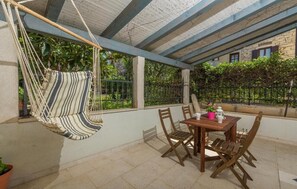 A covered patio with a wooden table and chairs, a hanging hammock, and a view of greenery and a stone building in the background. - Charming City-Center Cottage Just 20 Meters from the Beach in Novigrad (Novigrad)