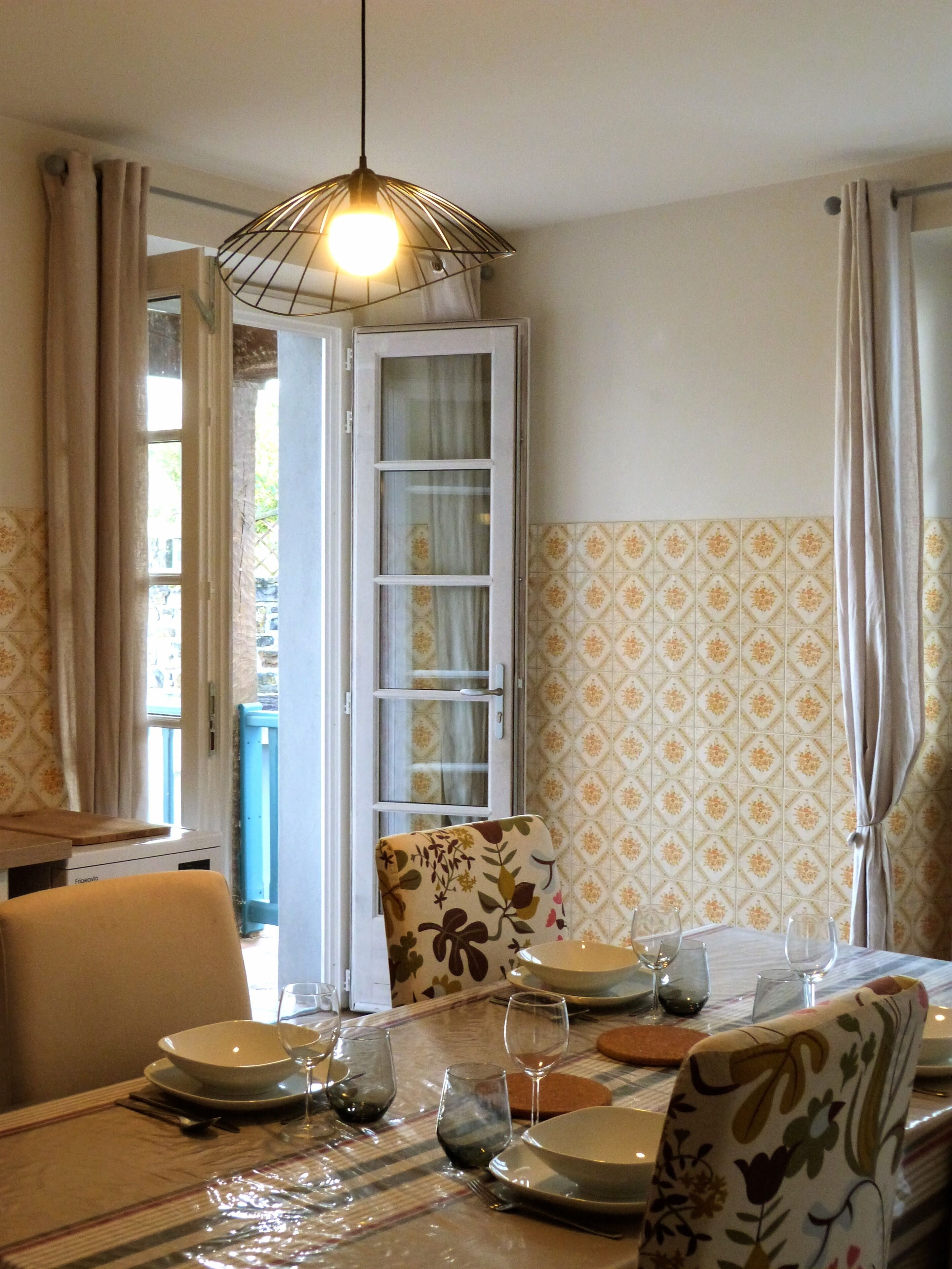 Un coin repas avec une table dressée pour deux personnes, une chaise à motifs floraux et un luminaire suspendu.