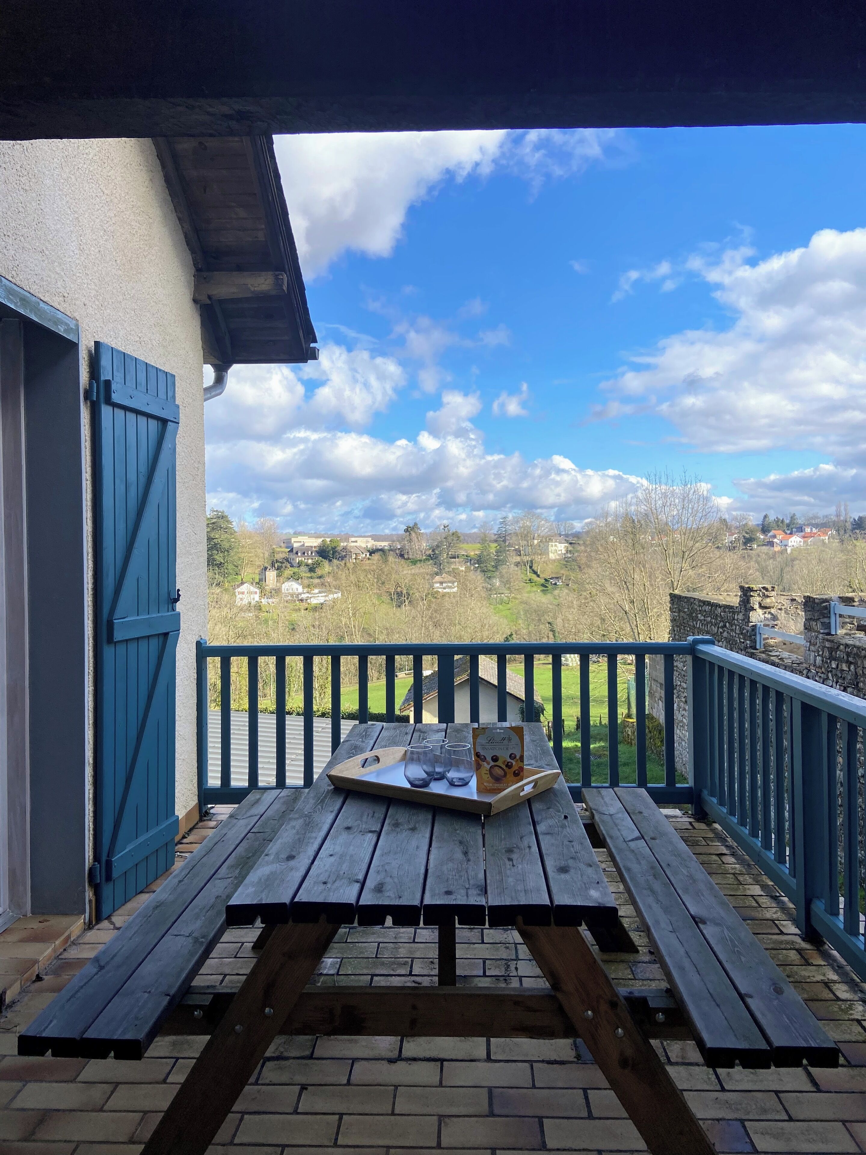 A wooden picnic table with a tray of glasses and a cookie box on a balcony with a blue railing.
