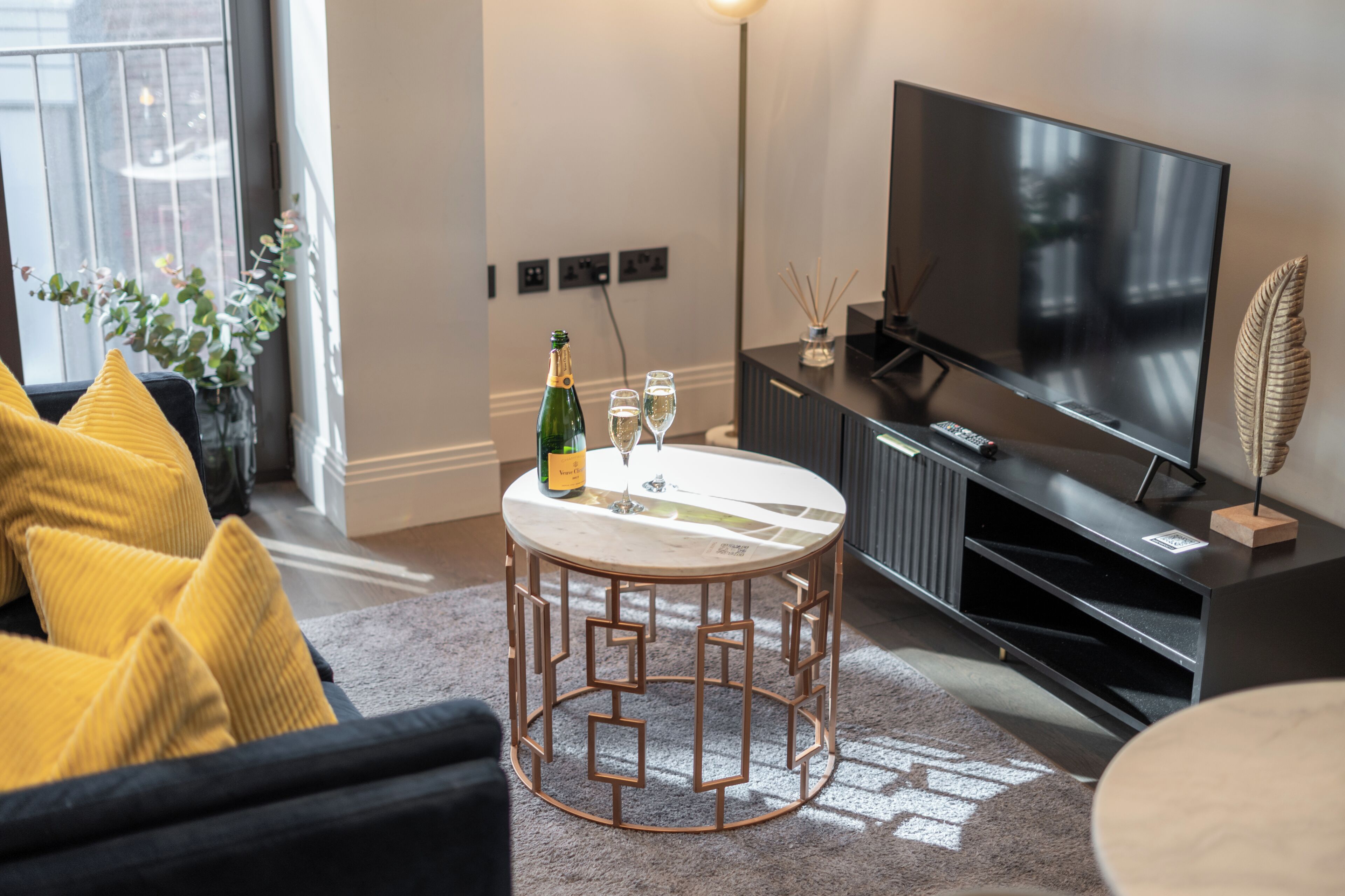 Un salon moderne avec une table basse ronde, un téléviseur à écran plat, un canapé avec des coussins jaunes et une plante en pot.