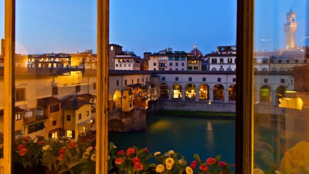 A view of a historic cityscape with buildings along a river, a prominent tower, and a bridge. - PONTE VECCHIO VIEW FLORENCE (Firenze)