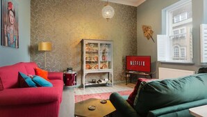 Ein Wohnzimmer mit einem roten Sofa, einem grünen Sofa, einem Schrank mit Geschirr, einem Netflix Fernseher und einem Fenster mit Blick auf ein Gebäude.