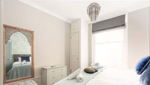 Ein Schlafzimmer mit einem großen Spiegel, einem Bett mit hellblau gestreifter Bettdecke, einem Nachttisch, einem kleinen Hocker und einer Kandelaberleuchte.