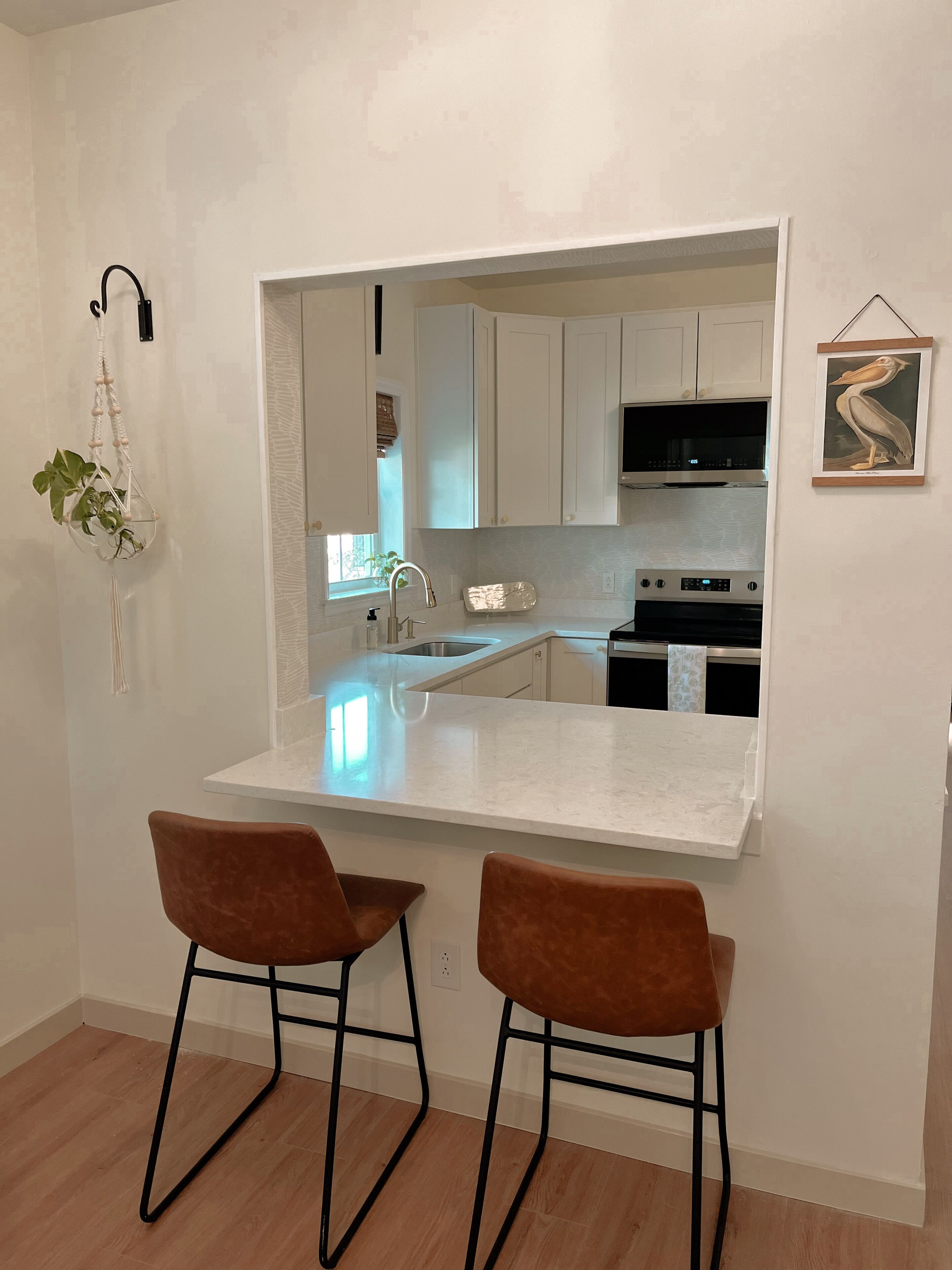 A modern kitchen with white cabinets, a large island, two brown bar stools, and a hanging plant.