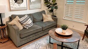 A living room with a grey sofa, two pillows, a round wooden coffee table with a plant, a large green plant beside the window, and two framed wall art pieces. - Beautifully renovated,  walkable home in Charleston - close to everything! (Charleston)