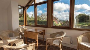 A sunlit room with wicker furniture, large windows, and a view of a rural landscape.