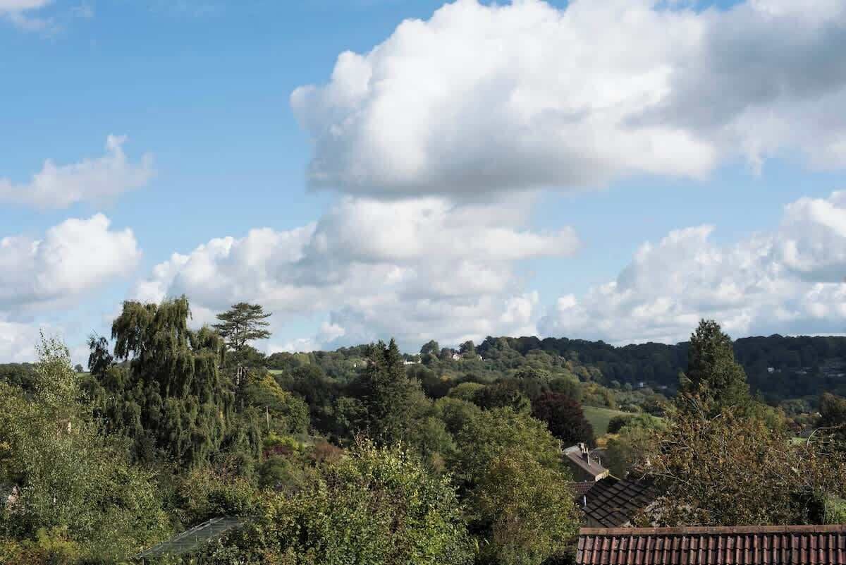A rural landscape with a house surrounded by trees and hills in the background.