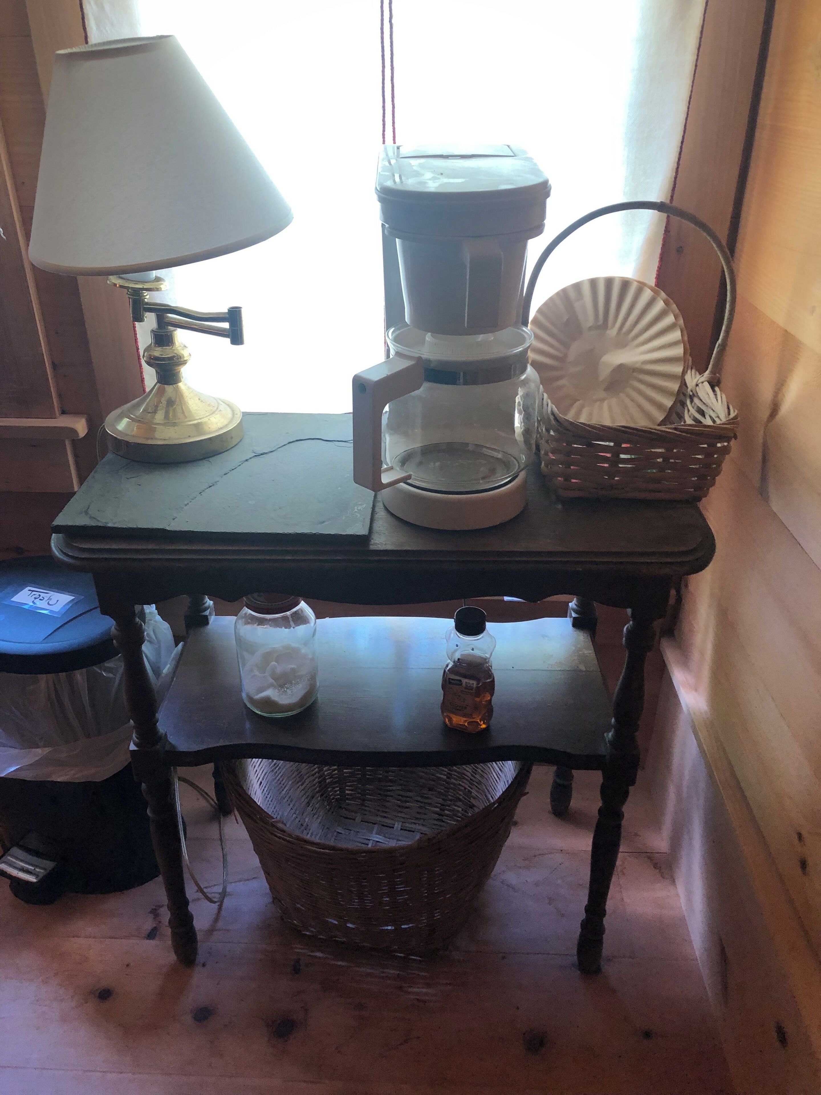 A wooden table with a coffee maker, a lamp, a jar, a honey bottle, and a basket.