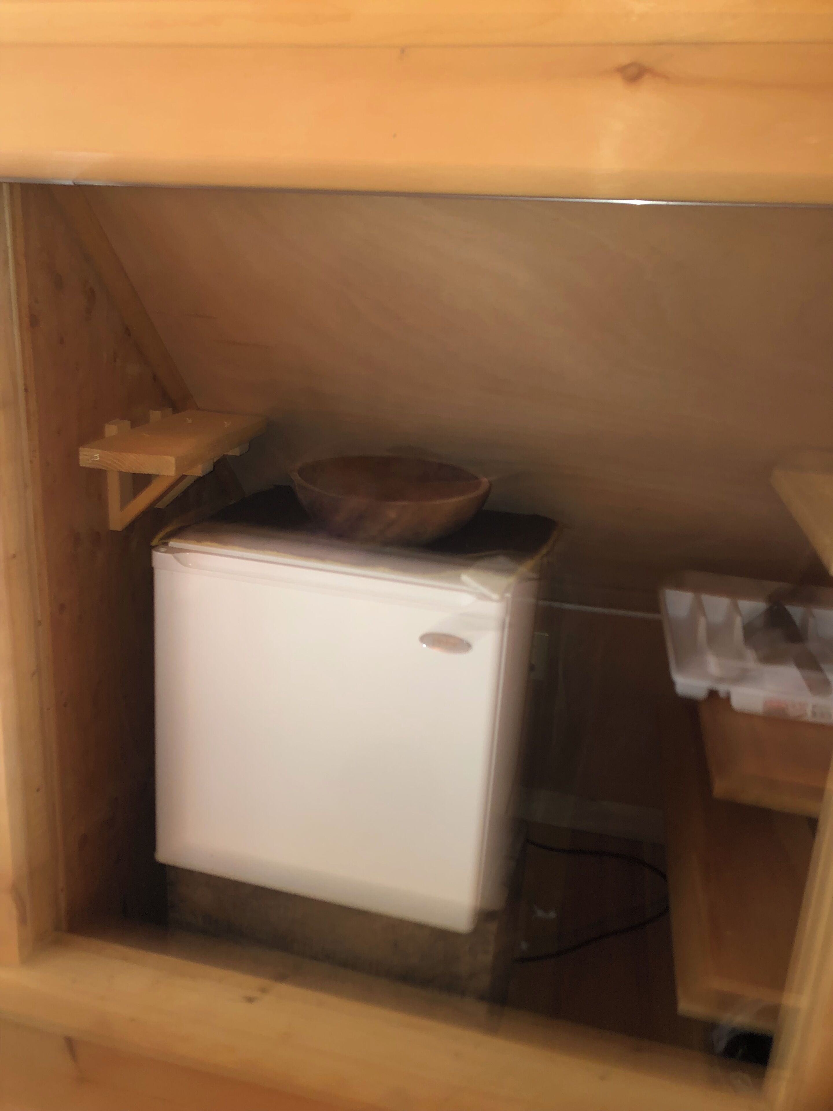 A compact kitchenette with a white refrigerator, a wooden bowl on top, and a small shelf.