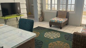 Una sala de estar acogedora con un televisor sobre un mueble bajo, una silla de mimbre, un banco acolchado y una alfombra estampada.