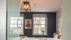 A bedroom with a bed, a window with a view of a building, a hanging light fixture, and a door with a glass panel.