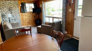 A cozy cabin interior with a stone wall, wooden ceiling, a dining table with chairs, a sofa, a television, and a fireplace.