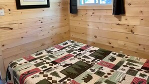 A cozy cabin room with wooden walls, a quilted bedspread, a framed picture, and a window with curtains.